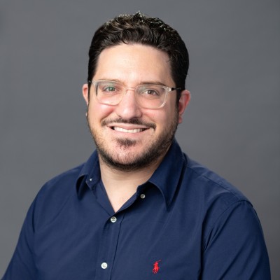 Aaron Goldman, wearing a blue shirt and glasses, smiles warmly at the camera.